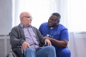 Male caregiver in blue scrubs talks with male senior patient in a wheelchair