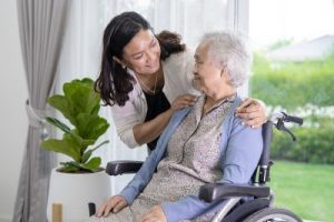 Young female caregiver embraces shoulders of senior female patient in a wheelchair